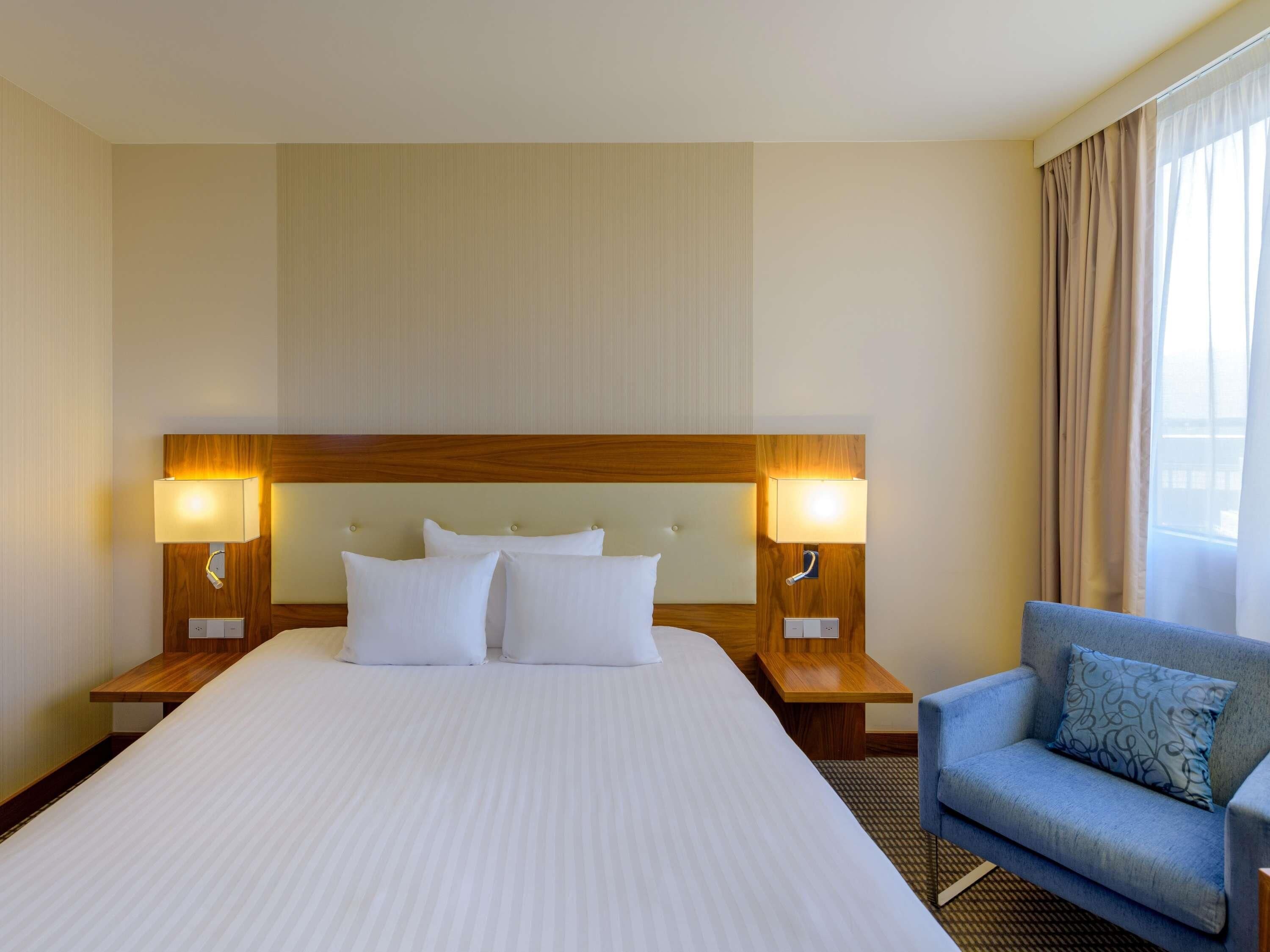 A hotel room with a large bed, two bedside lamps, a wooden headboard, a blue armchair, and a window with curtains.