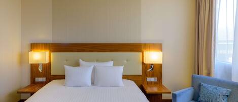 A hotel room with a large bed, two bedside lamps, a wooden headboard, a blue armchair, and a window with curtains.
