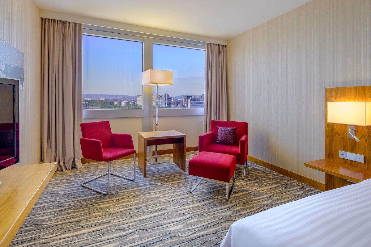 Habitación de hotel con dos sillones rojos, una mesita, una lámpara, una cama y un ventanal con vistas a la ciudad.