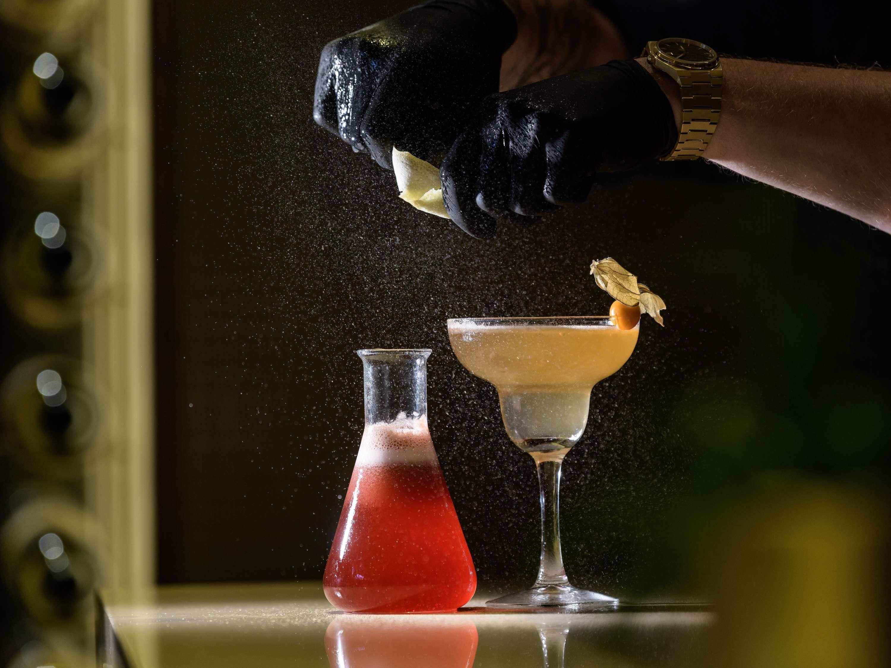 Une personne verse un liquide dans un verre à cocktail, avec une flasque et un autre cocktail sur une surface réfléchissante.