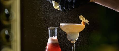 Une personne verse un liquide dans un verre à cocktail, avec une flasque et un autre cocktail sur une surface réfléchissante.