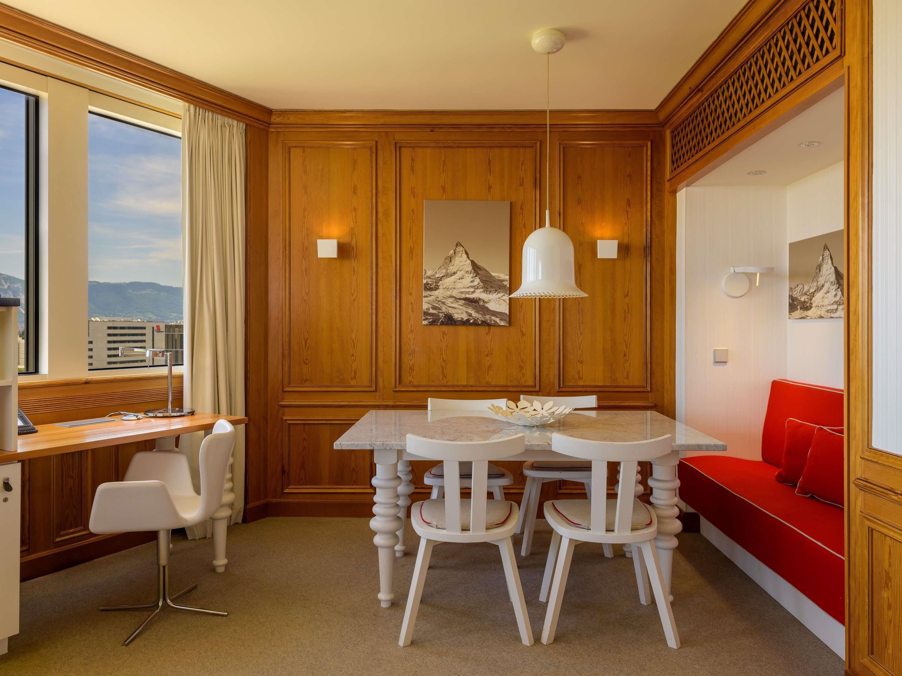 Quarto de hotel com parede de painéis de madeira, uma mesa de jantar com cadeiras, um sofá vermelho e uma janela grande com vista.