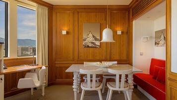 Quarto de hotel com parede de painéis de madeira, uma mesa de jantar com cadeiras, um sofá vermelho e uma janela grande com vista.