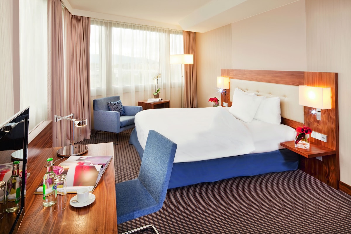 A hotel room with a large bed, a desk with a chair, a television, and a window with curtains.