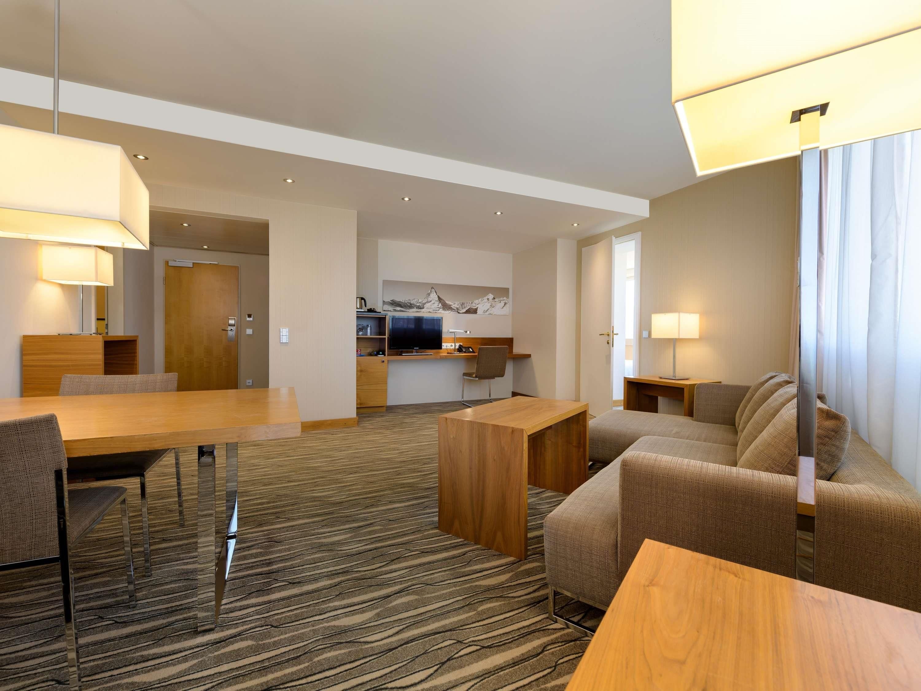 Ein modernes Hotelzimmer mit Essbereich, einer Sitzecke, einem Fernseher und einer Küche im Hintergrund.