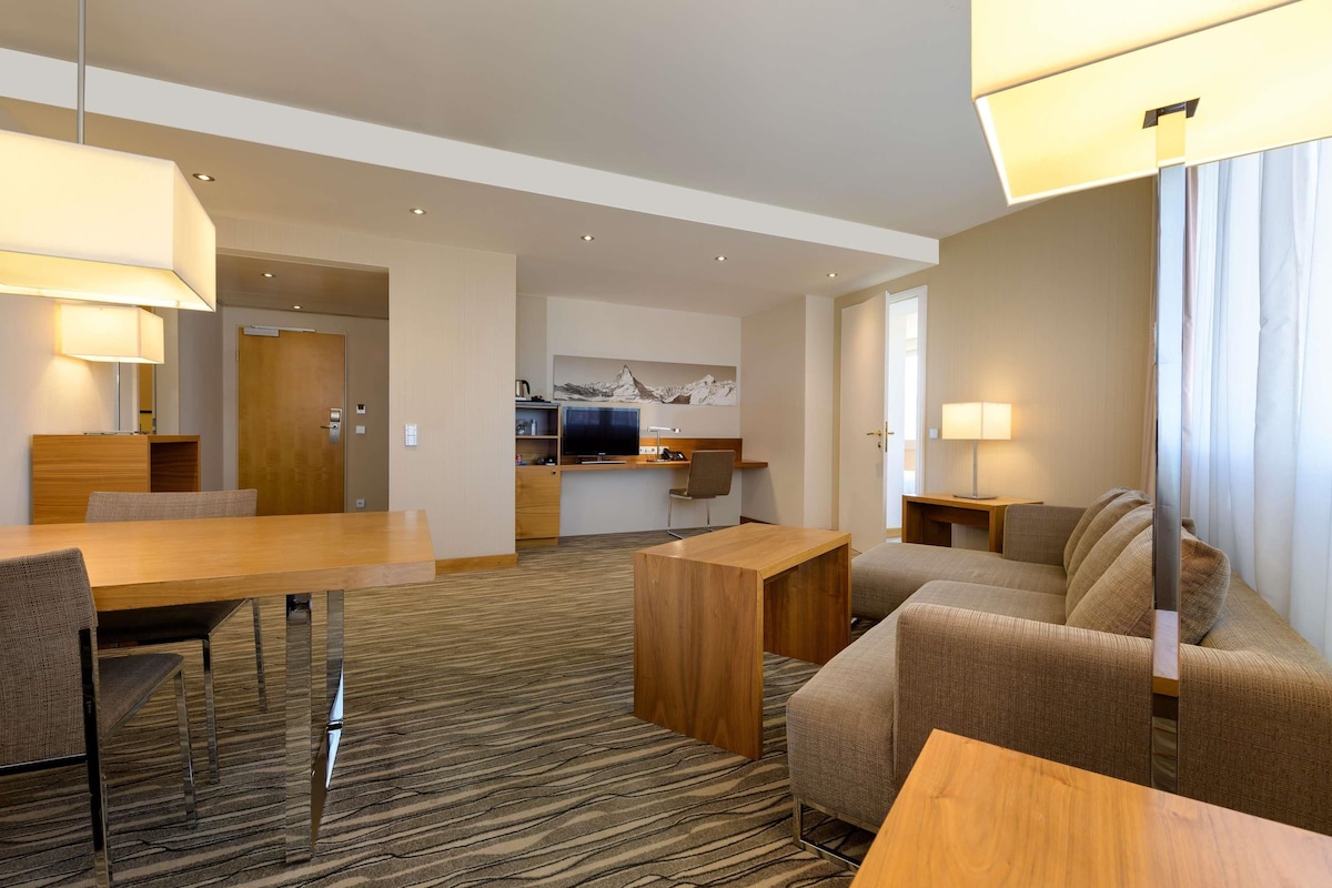 Una habitación de hotel moderna con comedor, sofá, televisión y una cocina al fondo.