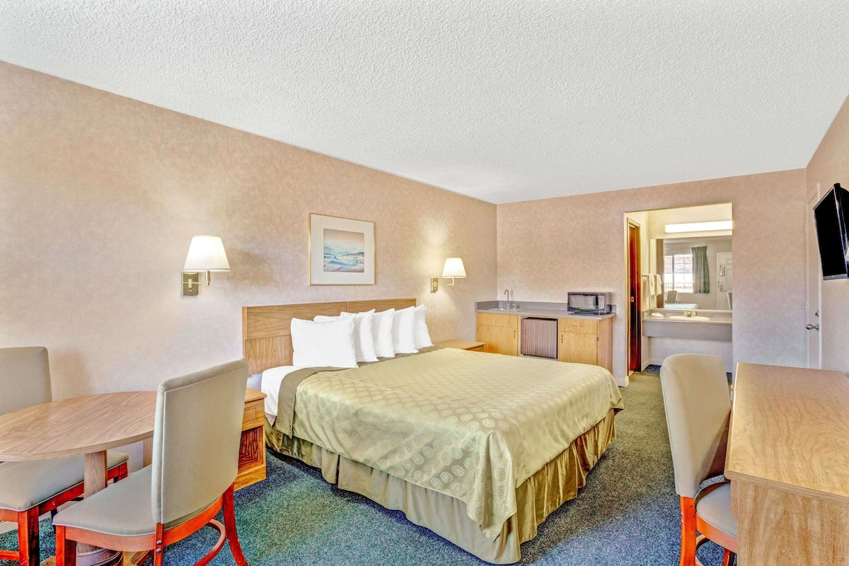 A hotel room with a bed, two bedside tables, a desk, a chair, a small kitchenette, and a bathroom visible through an open door.