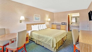 A hotel room with a bed, two bedside tables, a desk, a chair, a small kitchenette, and a bathroom visible through an open door.