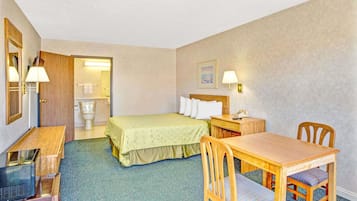 A hotel room with a bed, a desk, a chair, a table, a lamp, a mirror, a picture on the wall, and a bathroom with a toilet and sink.