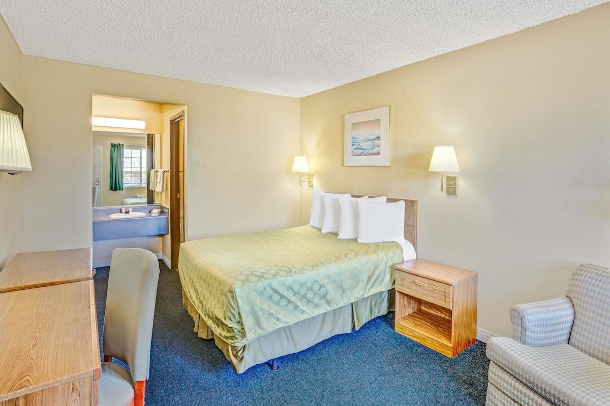 A hotel room with a bed, bedside table, chair, desk, and a bathroom visible through an open door.