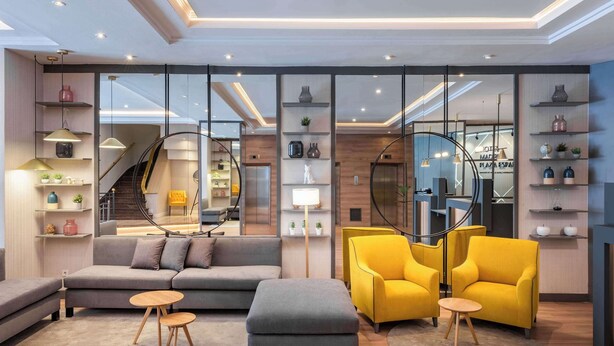 A modern living room with a sofa, ottoman, and yellow chairs, a staircase, and a glass partition with shelves. - Hotel Madrid Plaza de España Affiliated by Meliá (Madrid)