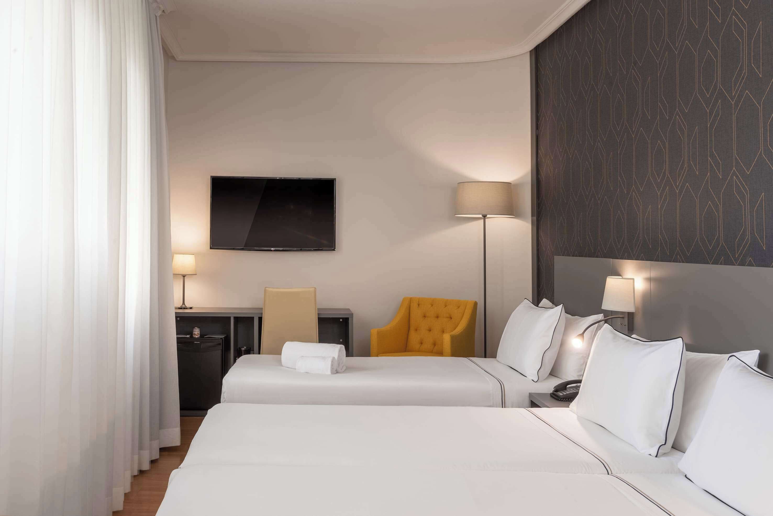 Une chambre d’hôtel avec deux lits, un téléviseur à écran plat, un bureau avec une lampe, une chaise et un mur décoré d’un motif.