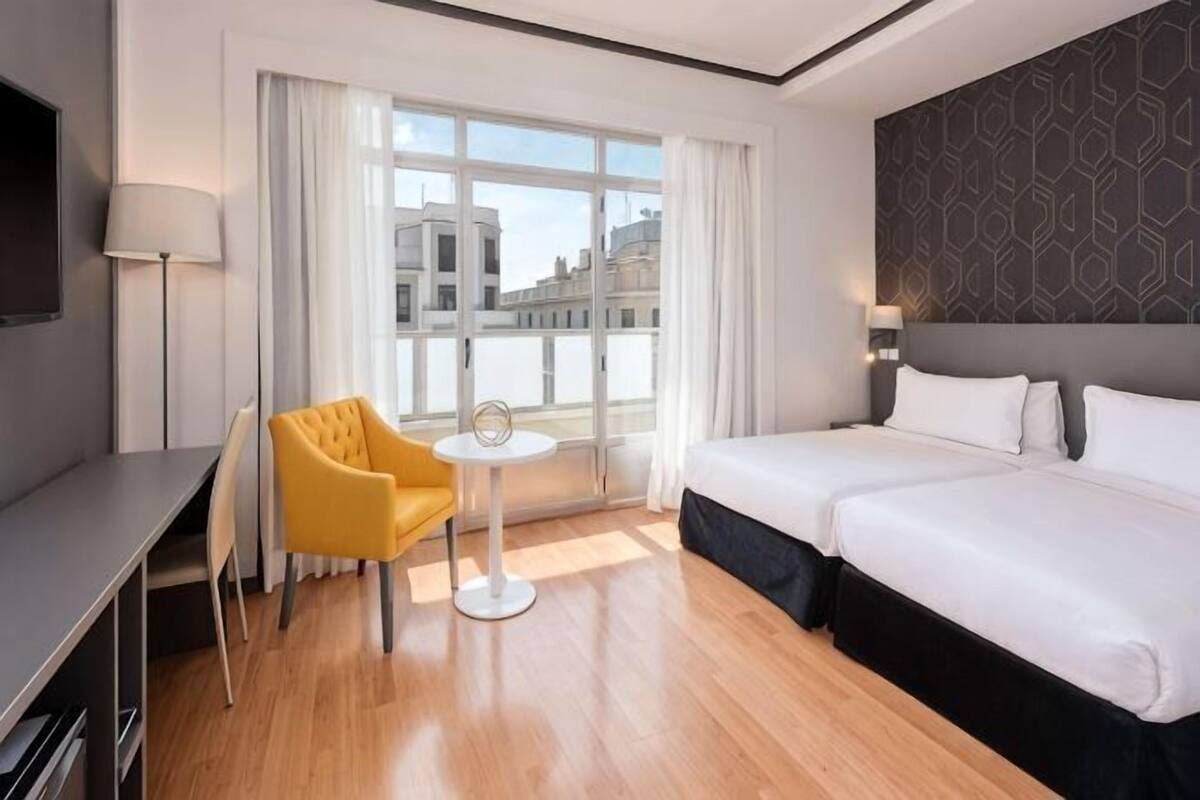 A hotel room with a bed, a desk, a chair, a small table, and a window with curtains.