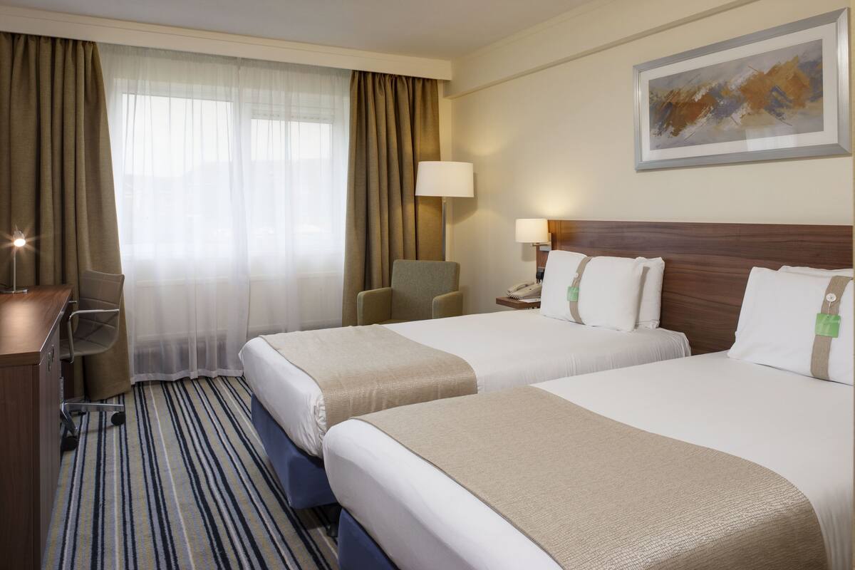 A hotel room with two beds, a desk, a chair, a lamp, and a painting on the wall.