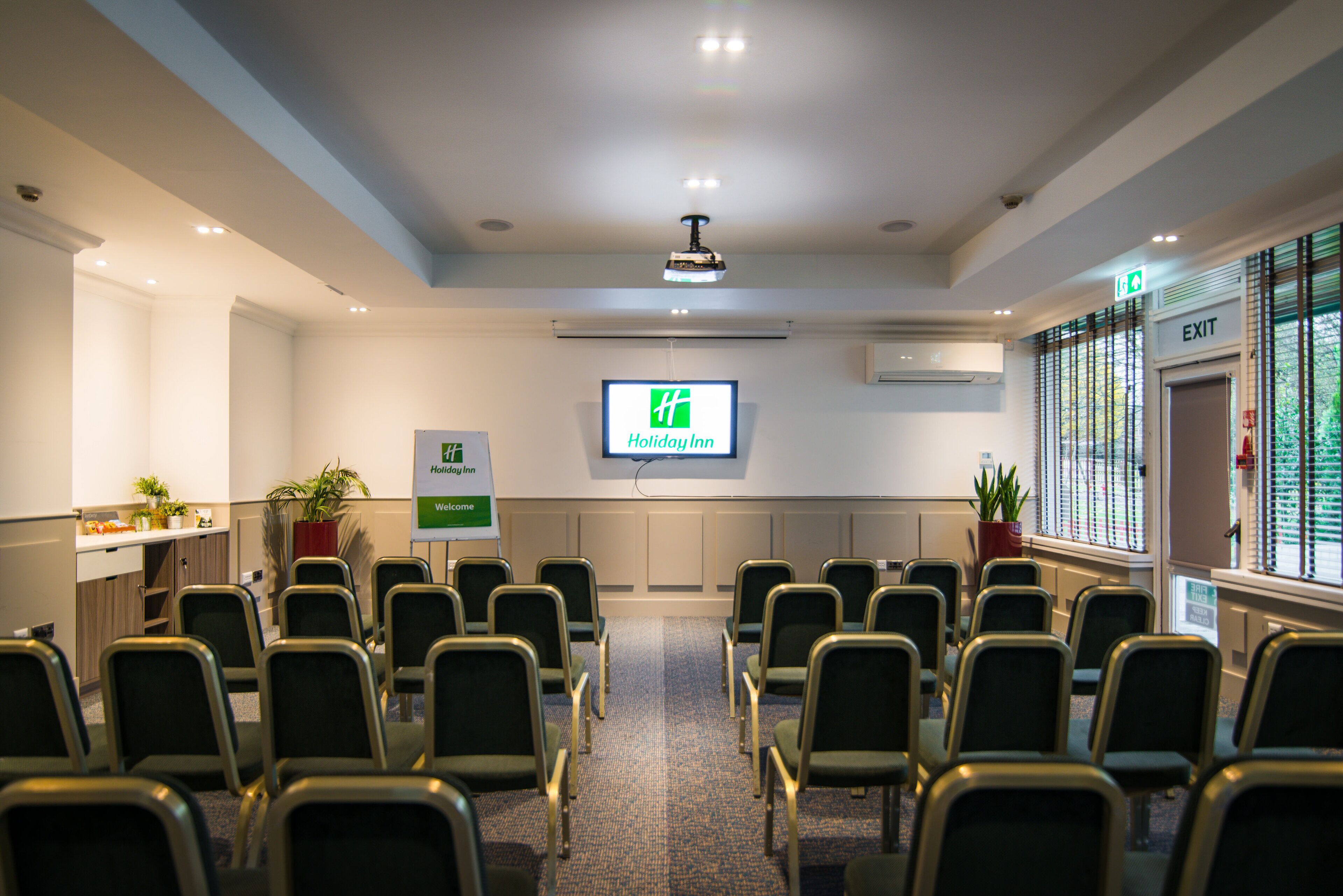 A conference room with rows of chairs, a screen displaying the Holiday Inn logo, and a reception area with plants and a counter.