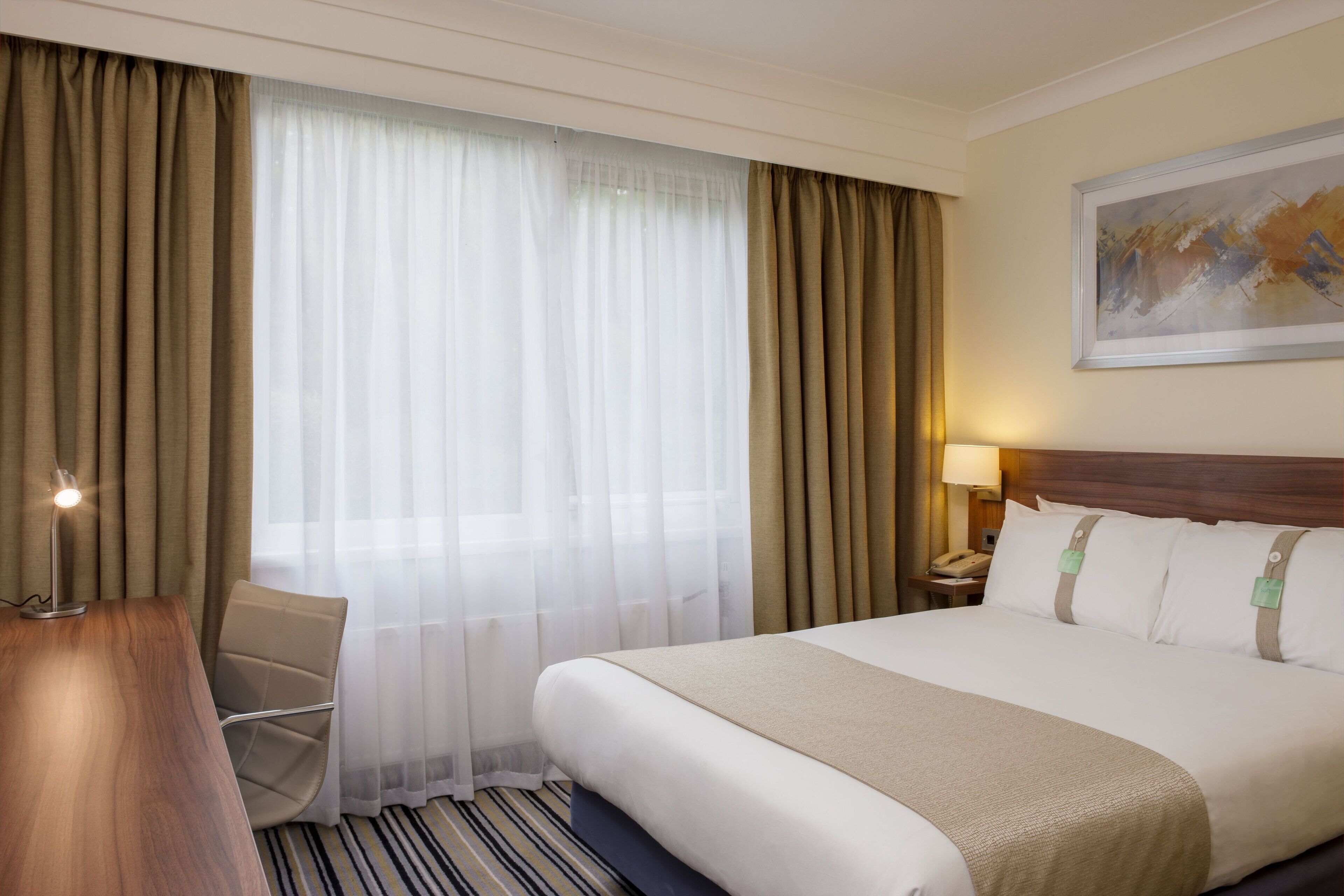 A hotel room with a large bed, a desk, a chair, a nightstand with a lamp, and a painting on the wall.