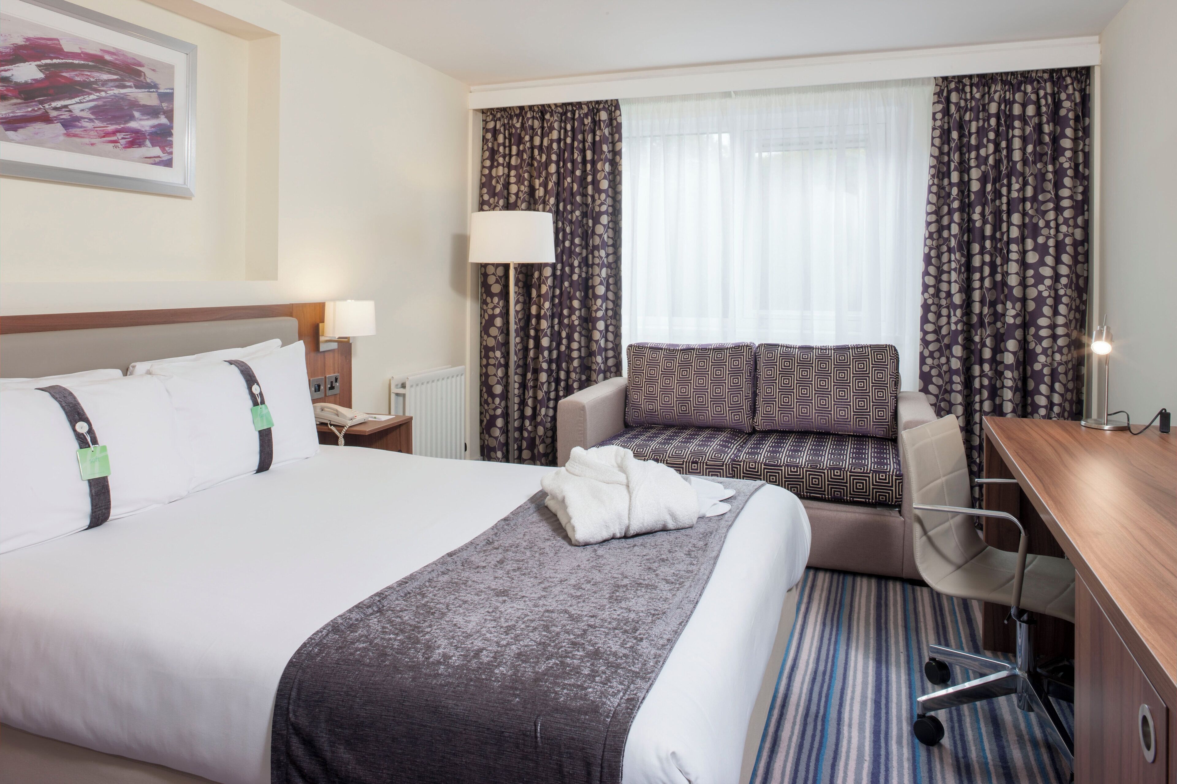 A hotel room with a large bed, a sofa, a desk, and a window with curtains.