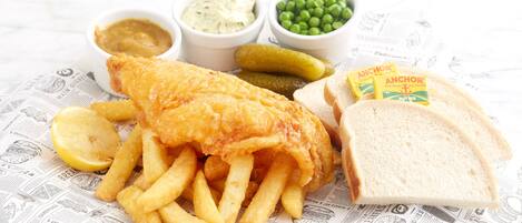 Un plato de pescado y papas fritas, acompañado de pepinillos, guisantes y salsas, servido sobre papel de periódico.