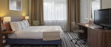 Habitación de hotel con una cama grande, un escritorio con televisor, una silla y una ventana con cortinas.