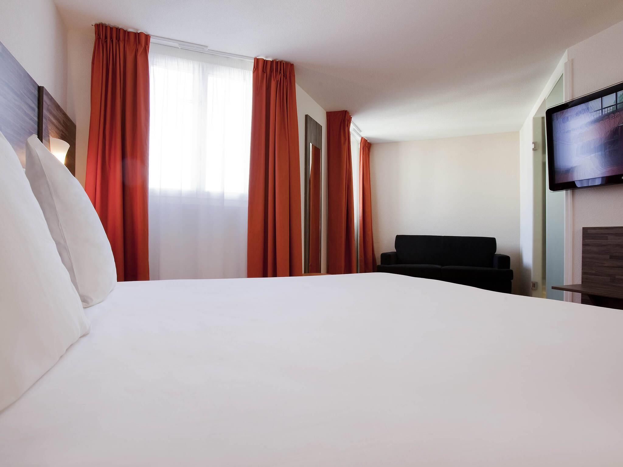A hotel room with a large bed, a black sofa, a flat-screen TV mounted on the wall, and red curtains on the windows.