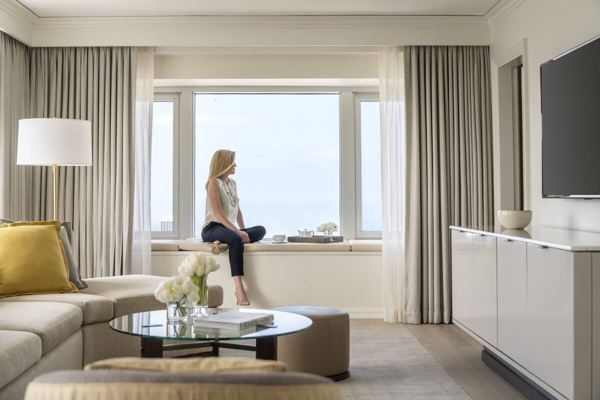 A modern hotel room with a large window, a sofa, a coffee table with flowers, and a TV on the wall.