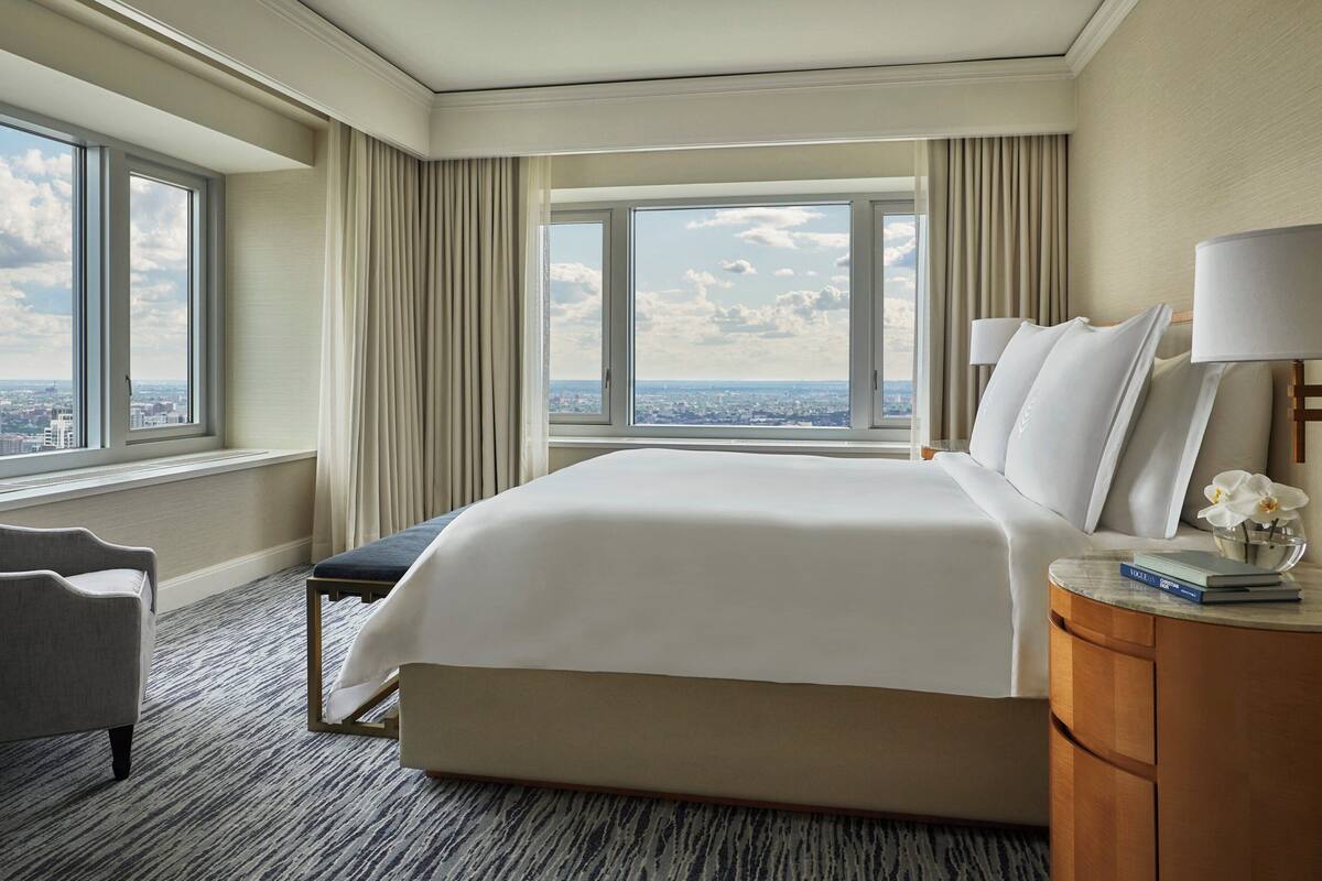 A hotel room with a large bed, a chair, a nightstand, and a view of the city through large windows.