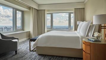 A hotel room with a large bed, a chair, a nightstand, and a view of the city through large windows.