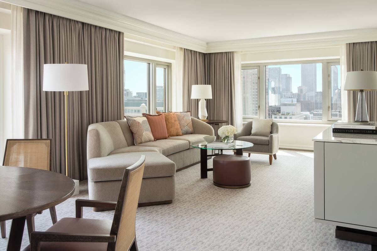 A modern hotel room with a sectional sofa, armchairs, a dining table, and a view of the city skyline.