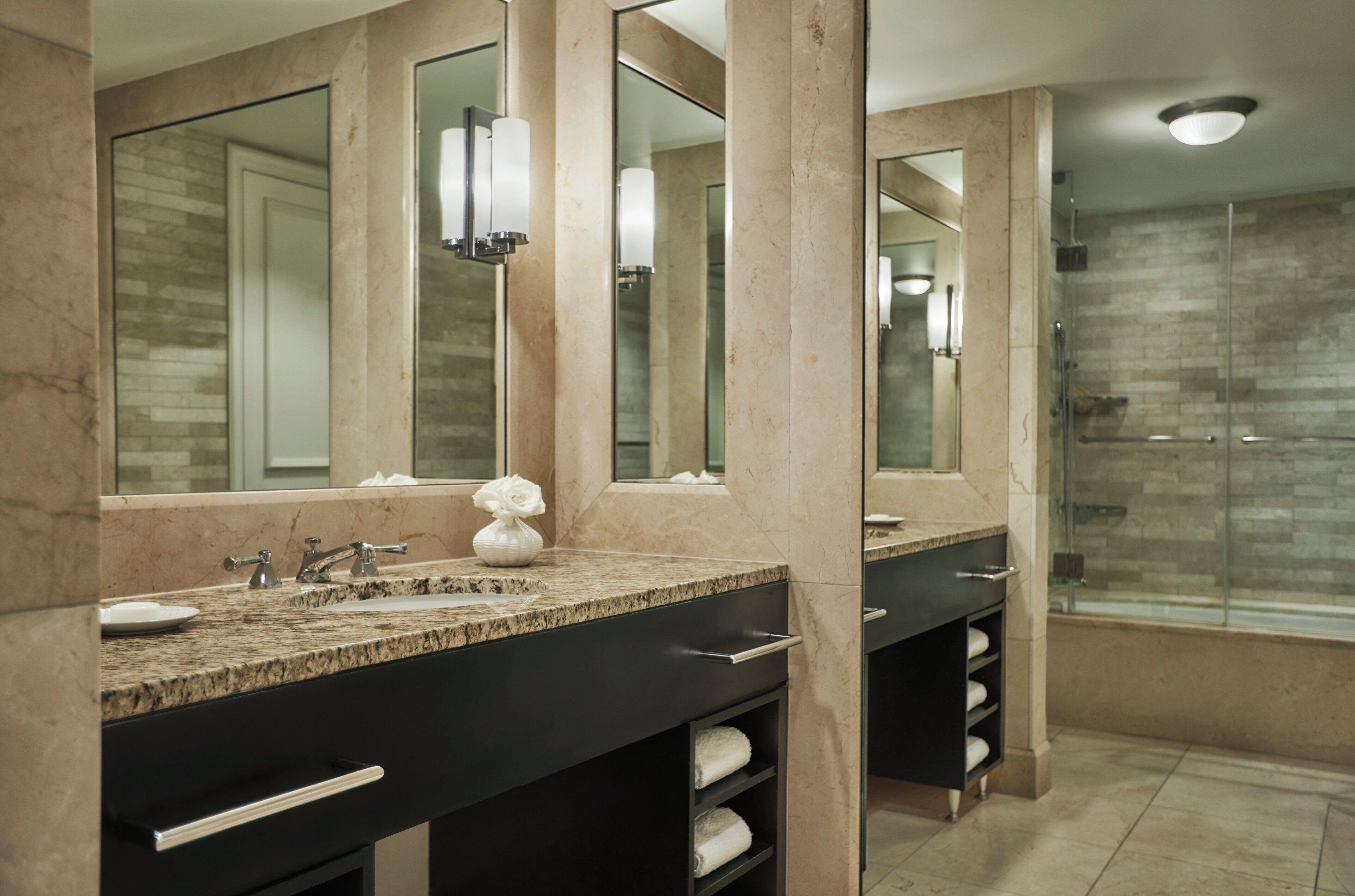 A modern bathroom with a granite countertop, a large mirror, a glass shower enclosure, and a marble wall.