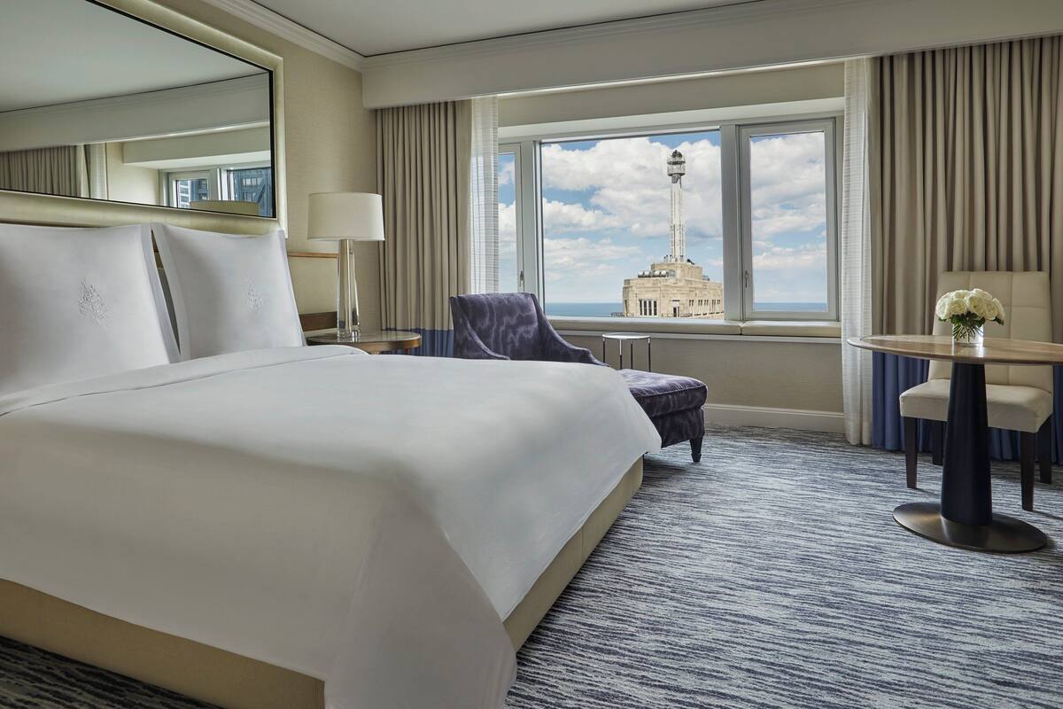 A hotel room with a large bed, a chair, a table, and a view of a tower through a window.