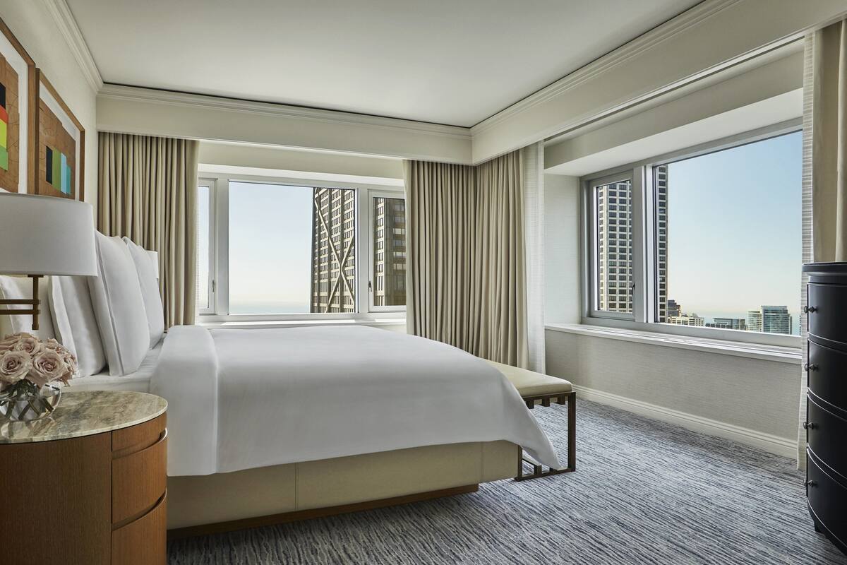 A hotel room with a large bed, a bedside table with a lamp, a vase of flowers, and a view of the city through large windows.