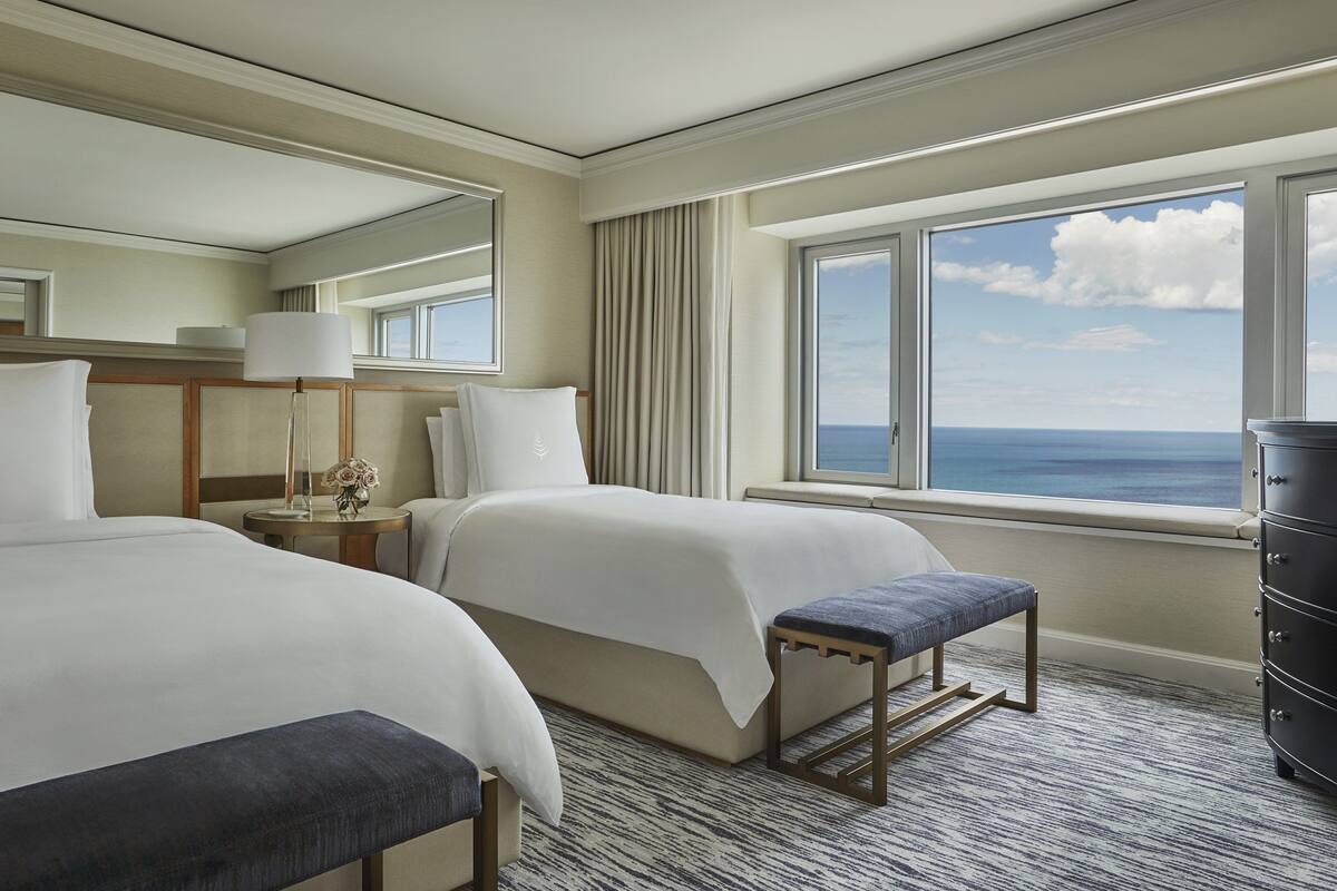 A hotel room with two beds, a large window with a view of the ocean, a bedside table with a lamp, and a dresser.