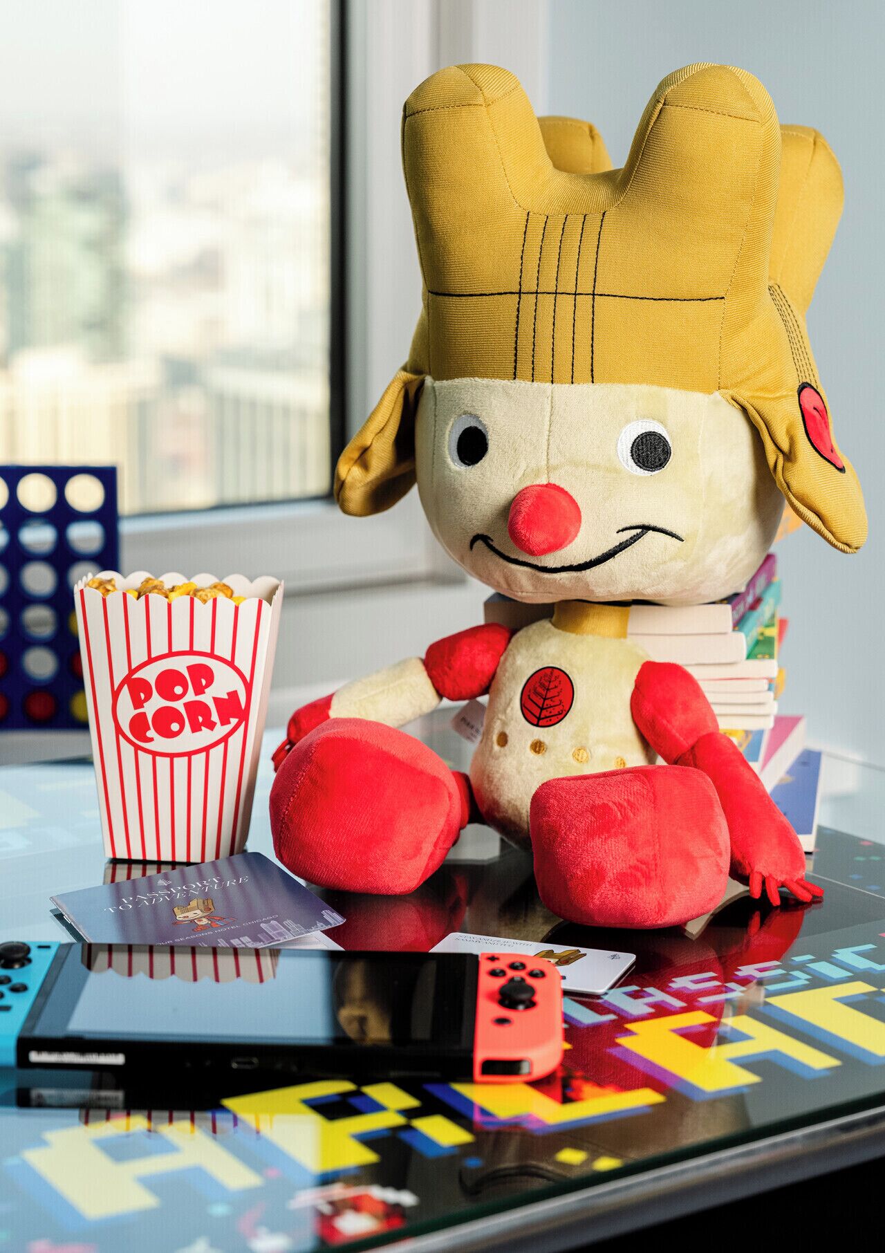 Un jouet en peluche avec une couronne, un récipient à pop-corn et une pile de livres, le tout posé sur une surface réfléchissante.