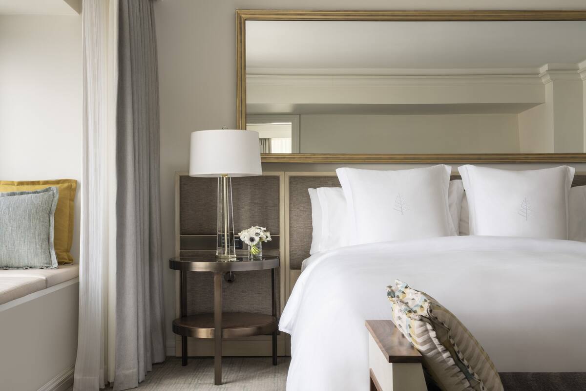 A hotel room with a large bed, a bedside table with a lamp, a mirror, and a window with curtains.