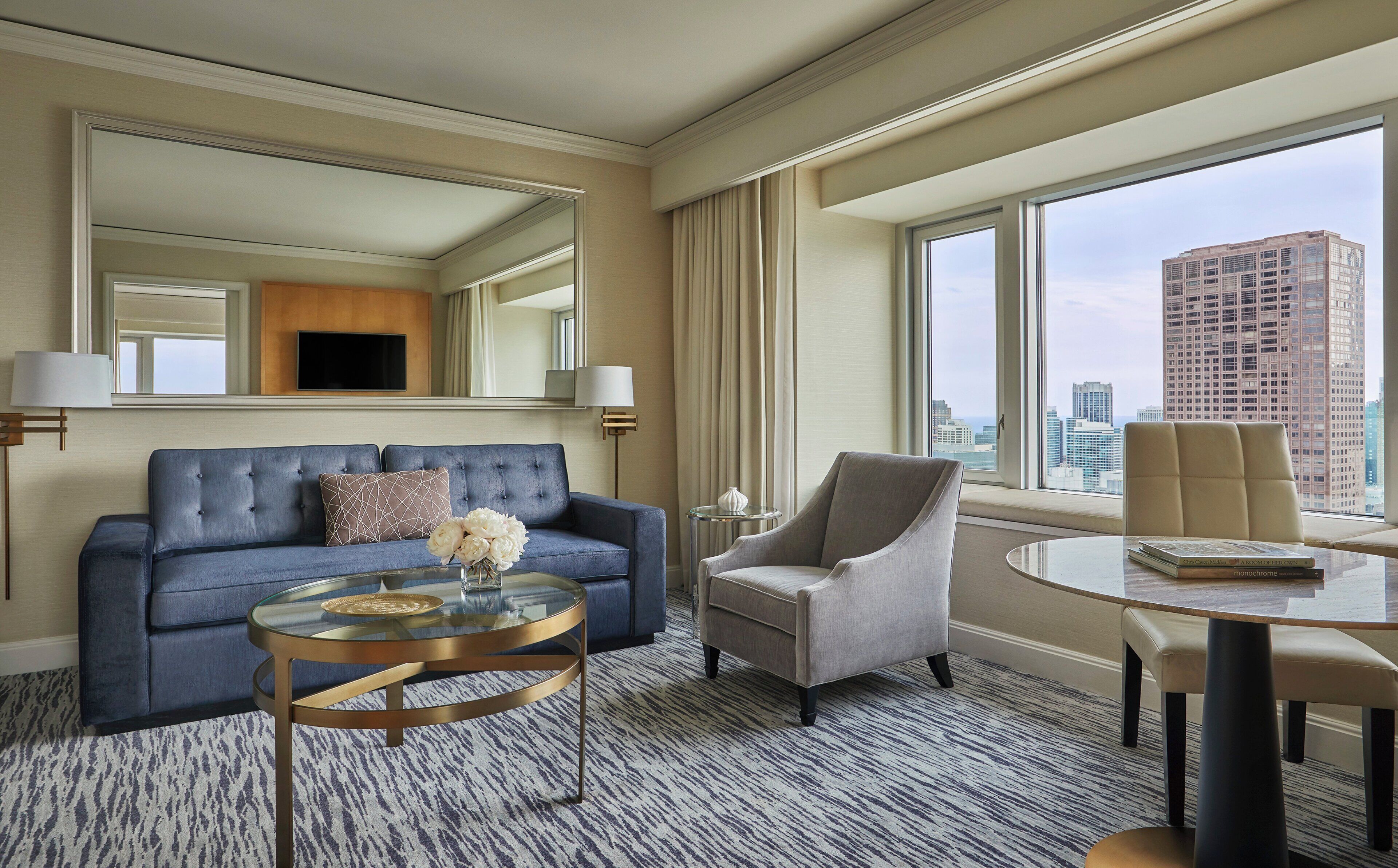 Habitación de hotel moderna con un sofá azul, un sillón gris, una mesa de centro de vidrio y vistas a la ciudad a través de amplias ventanas.
