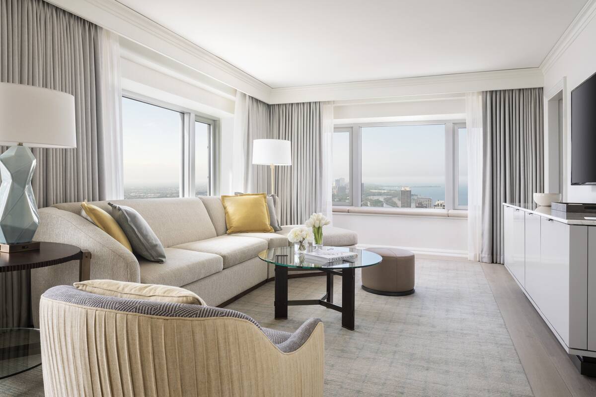 A modern hotel room with a sofa, armchair, glass coffee table, and a view of the city through large windows.