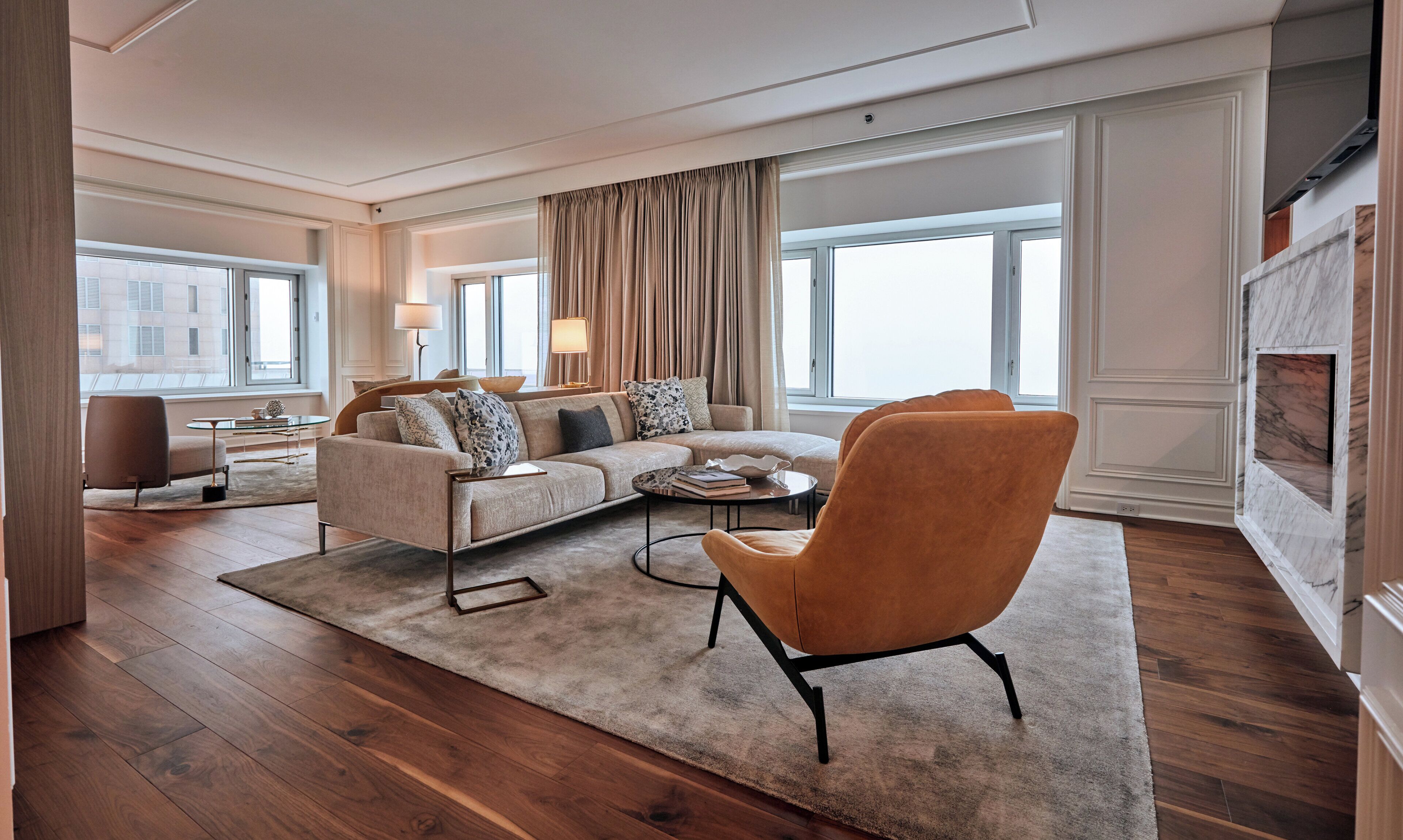 A modern living room with a sectional sofa, a round coffee table, and a single armchair. There is a large window with curtains, a small side table with a lamp, and a view of the cityscape.