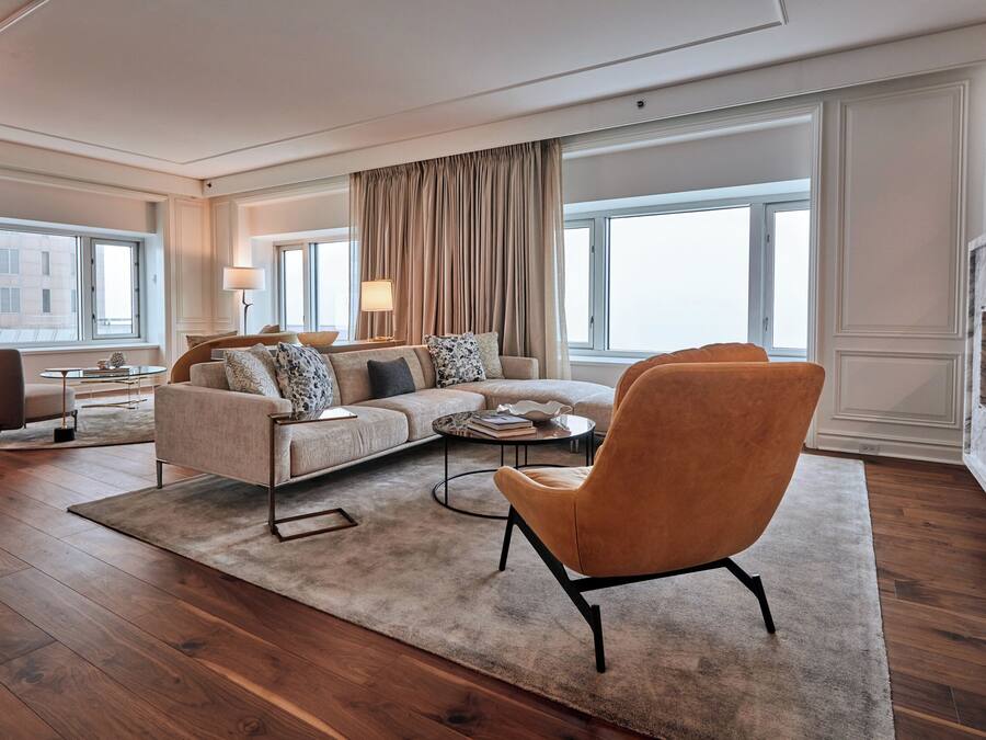 A modern living room with a sectional sofa, a round coffee table, and a single armchair. There is a large window with curtains, a small side table with a lamp, and a view of the cityscape.