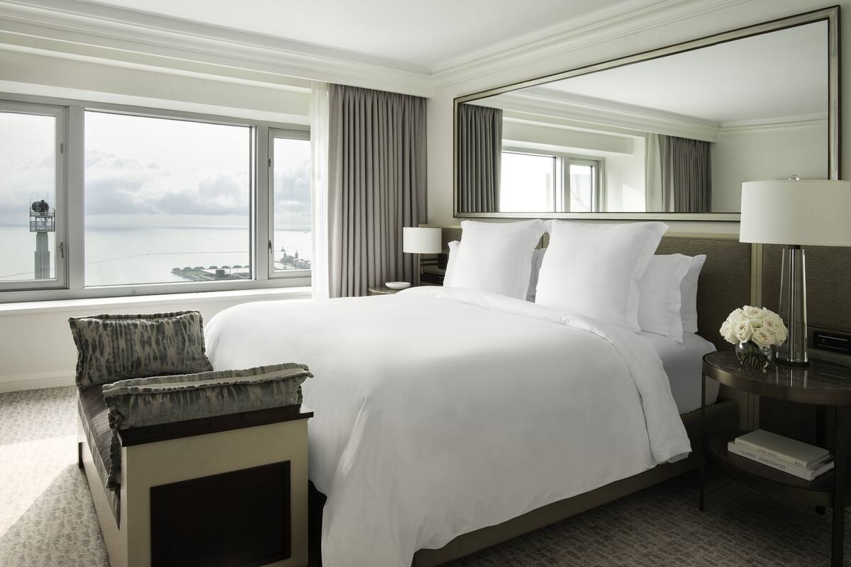 A hotel room with a large bed, a bedside table with a lamp, a chair, a large window with a view of the sea, and a mirror on the wall.