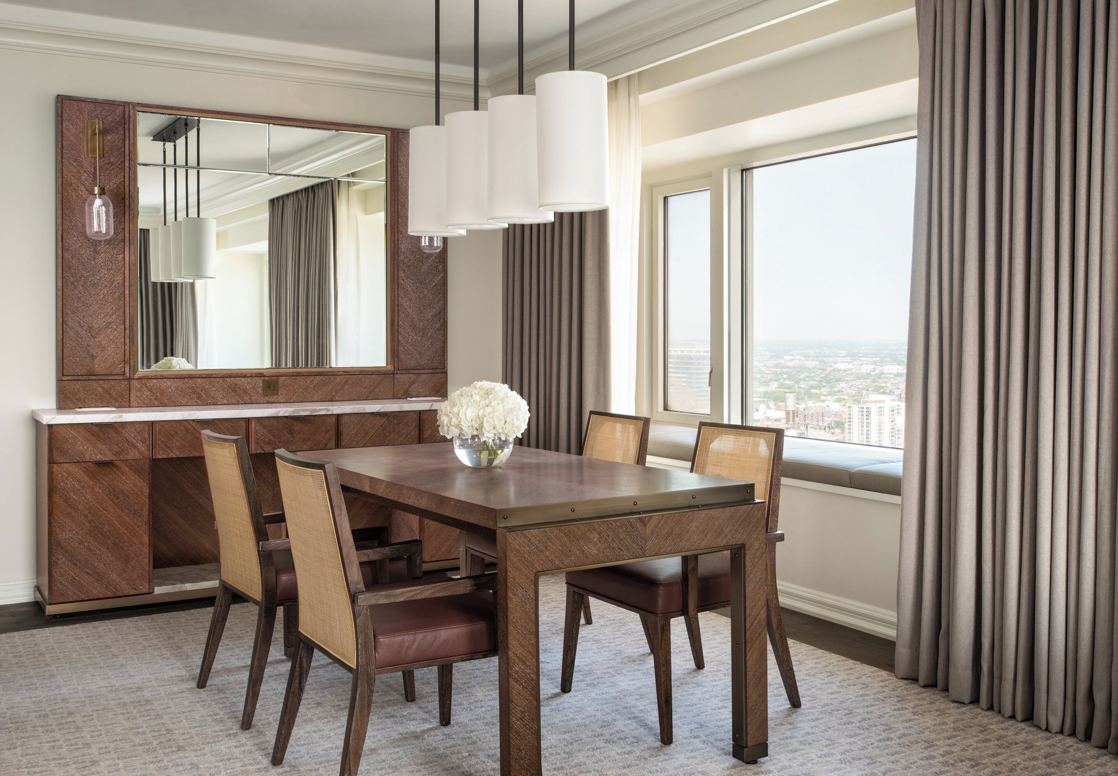 Ein modernes Hotelzimmer mit Essbereich, ausgestattet mit einem Holztisch und Stühlen, einem großen Spiegel und Blick auf die Stadt durch große Fenster.