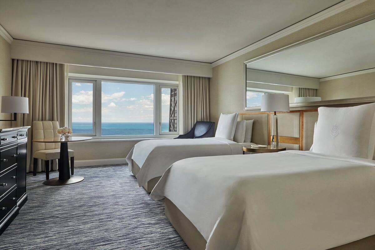 A hotel room with two beds, a desk, a chair, a lamp, and a large window with a view of the ocean.