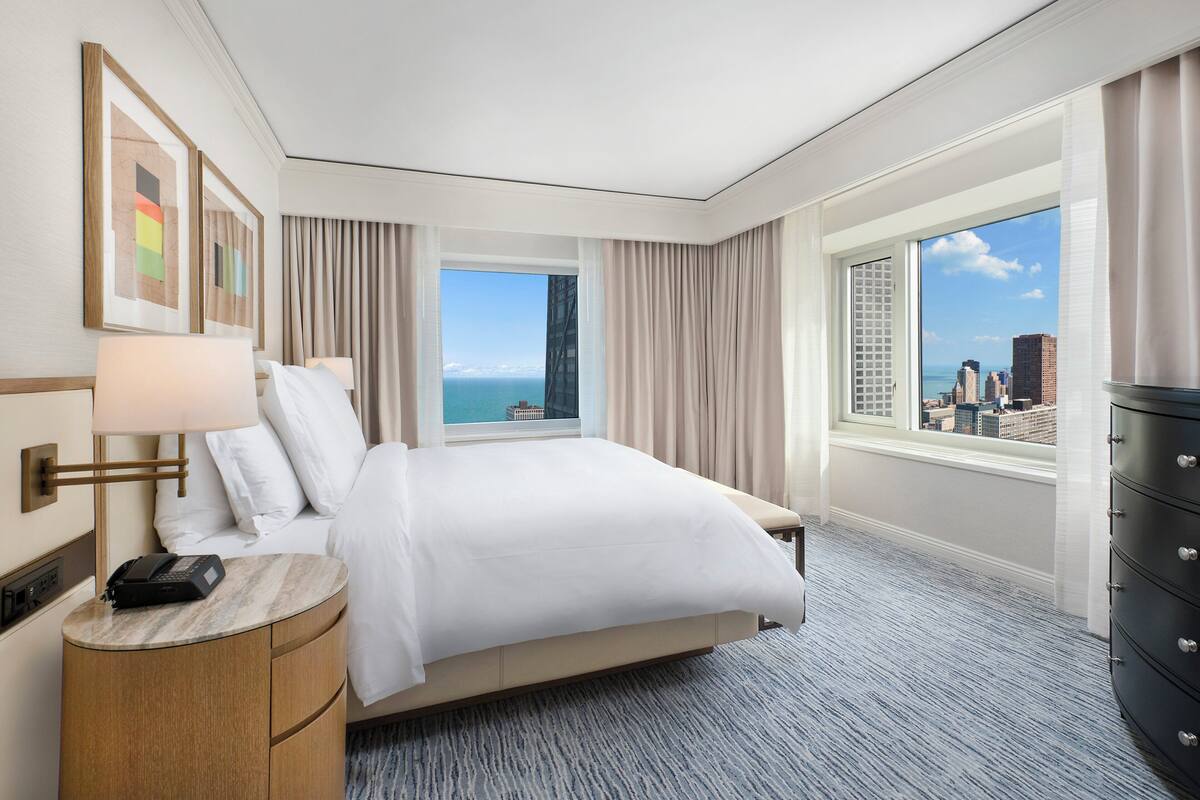 A hotel room with a bed, a desk, a dresser, and a large window with a city view.