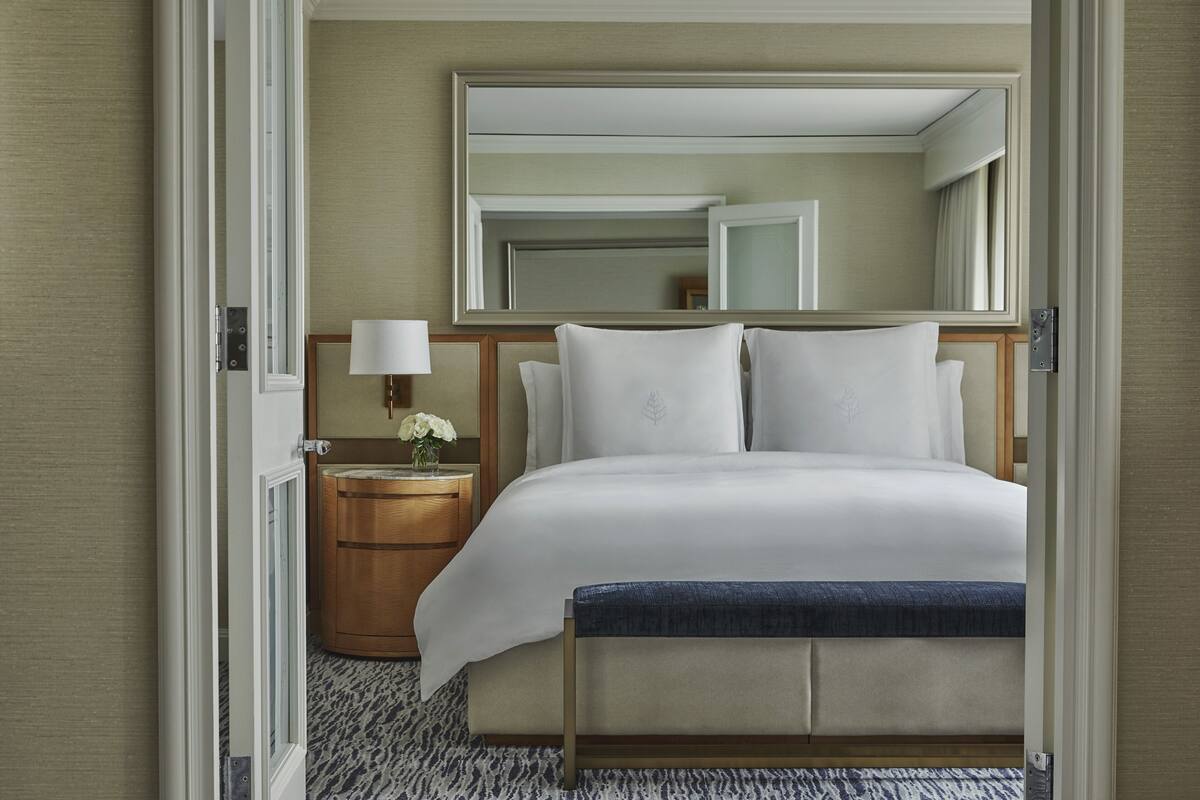 A hotel room with a large bed, a bedside table with a lamp, a mirror above the bed, and a wooden nightstand with a vase of flowers.