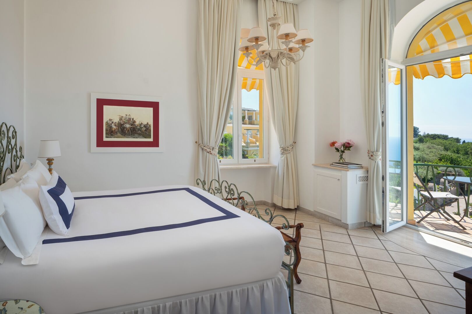 Un dormitorio con cama, un cuadro en la pared, una ventana con cortinas, una araña y un balcón con mobiliario exterior.
