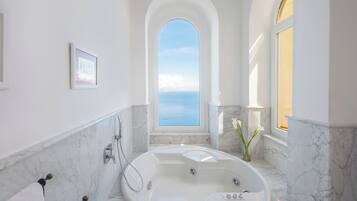 Un baño moderno con una bañera grande, paredes de mármol y vista al mar a través de un ventanal de arco.