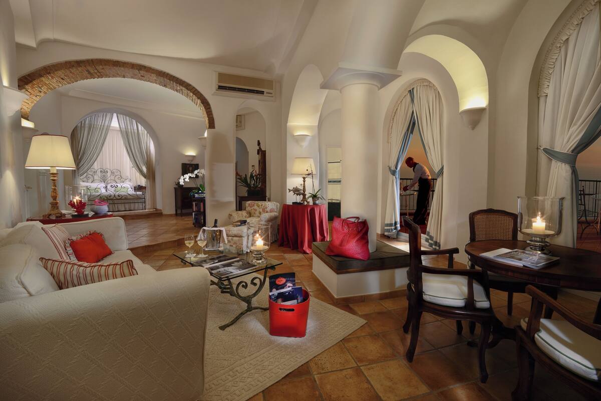 A spacious living room with a sofa, armchairs, a coffee table, and a dining table. There are decorative lamps, a red handbag, and a vase with flowers.