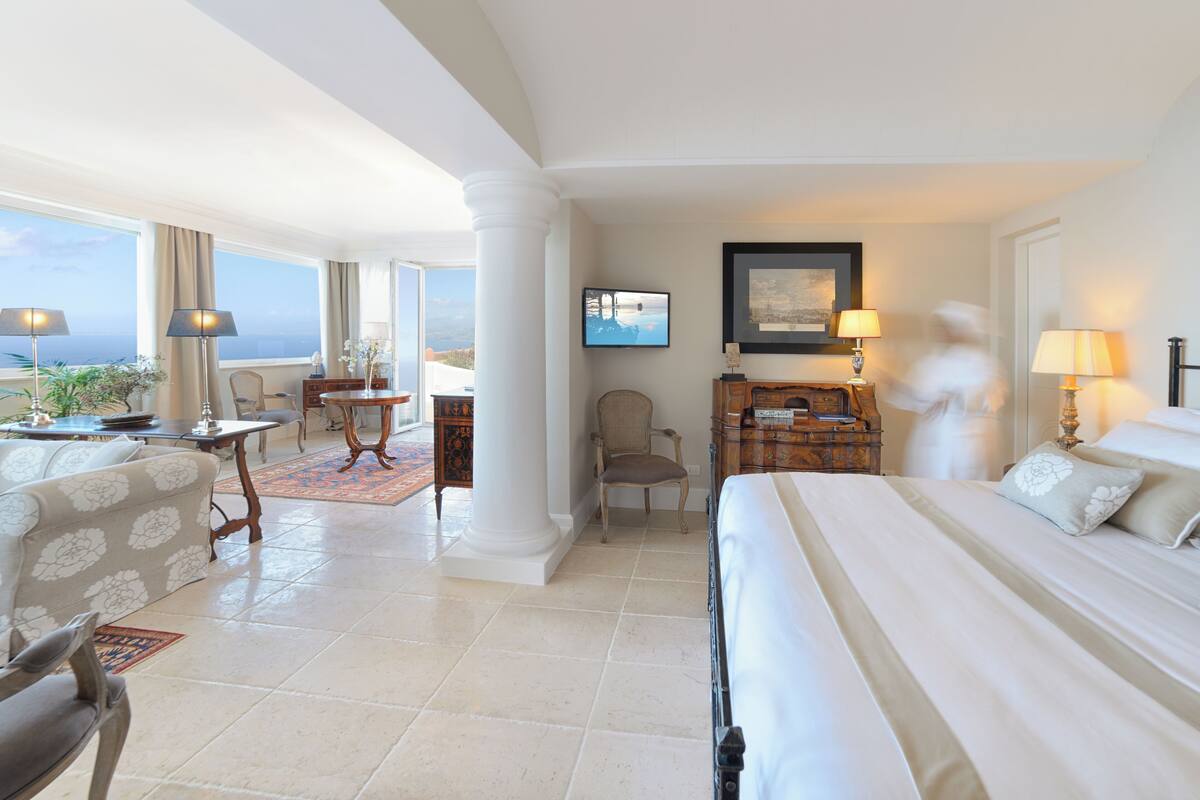 A hotel room with a large bed, a desk, a chair, and a television. There is a view of the ocean through the windows.