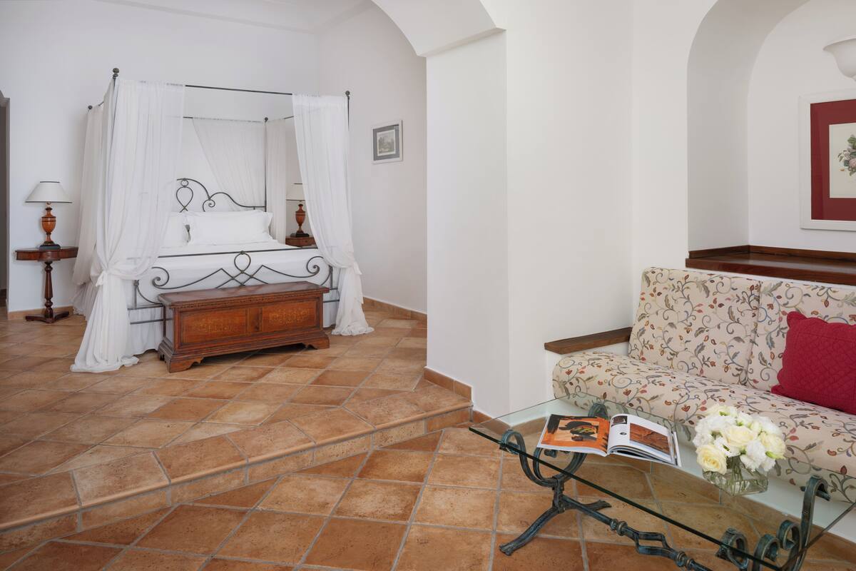 A bedroom with a four-poster bed, a wooden dresser, a sofa with a floral pattern, and a glass coffee table with a book and flowers.