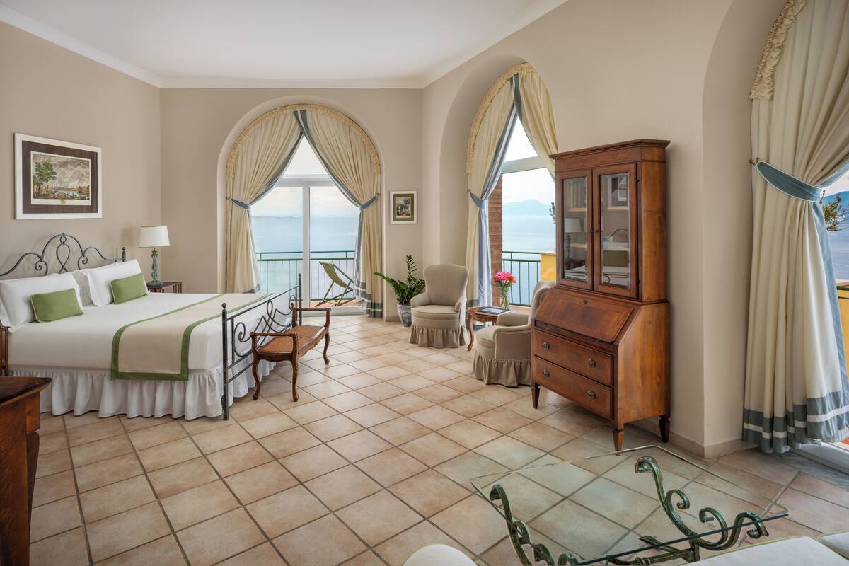 A hotel room with a bed, a wooden cabinet, a chair, a table, and large windows with curtains.