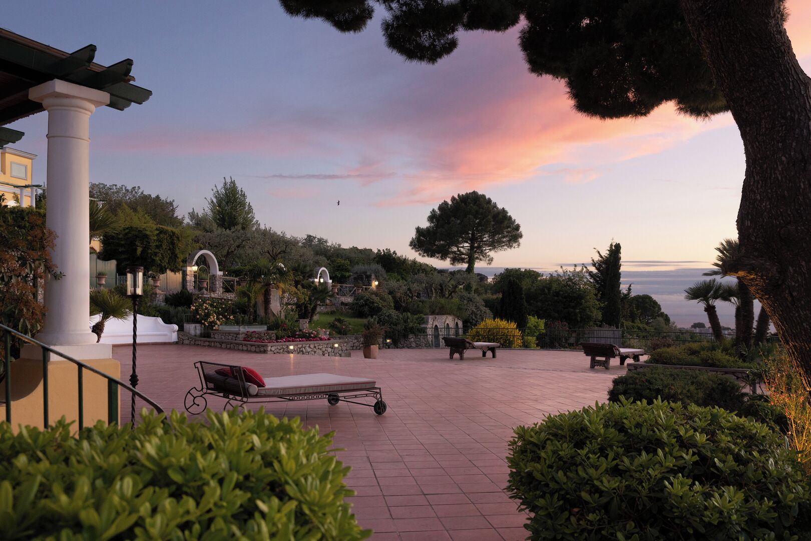 A courtyard with a sun lounger, benches, and a view of the sea at sunset.